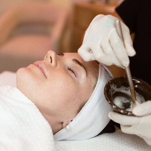 A person wearing a white headband lies down while a gloved professional prepares a treatment with a brush and bowl.