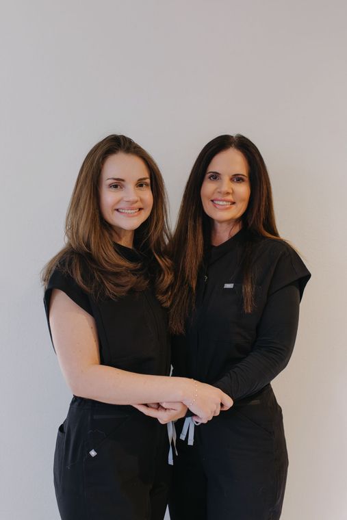 Two women in black scrubs smiling, standing together against a white wall.