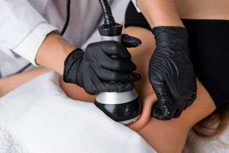 A practitioner wearing black gloves uses a handheld ultrasonic device on a client’s thigh during a spa treatment.