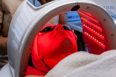 A person wearing protective goggles lies under a curved LED device emitting bright red light during a facial treatment.