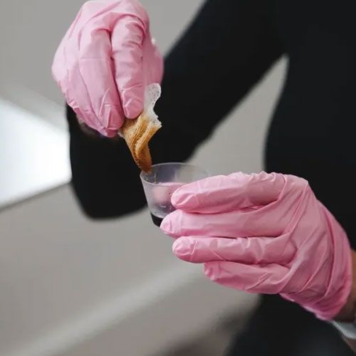 A person wearing pink gloves dips a gauze pad into a small medicine cup.
