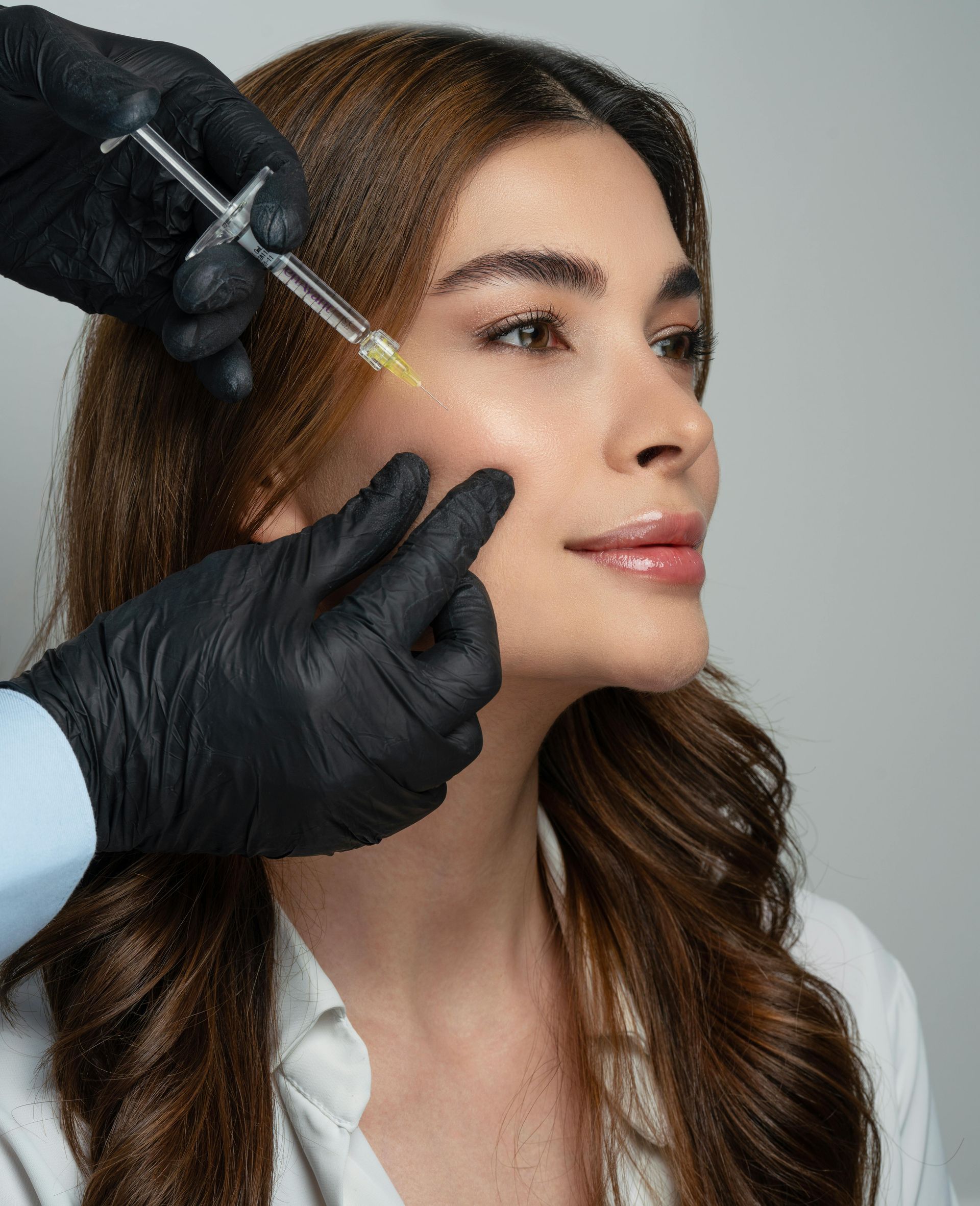 Person receiving a facial injection. Hand in black glove holding syringe near cheek. Woman with brown hair.