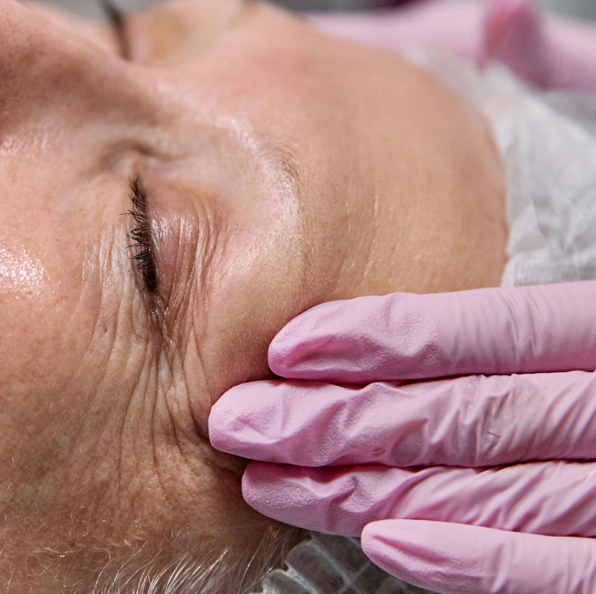 A person with short, gray-streaked hair, eyes closed, gently touching their cheek against a solid tan background.