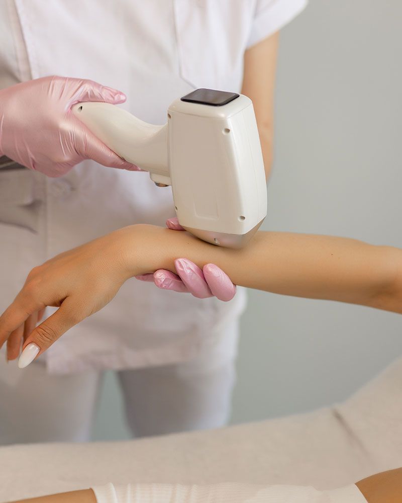 Person using a light-based device on a forearm for hair removal, wearing gloves and a white uniform.