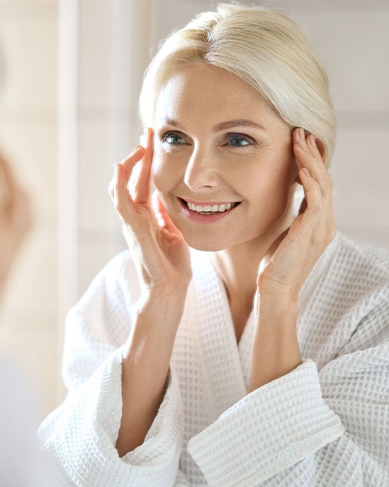 Woman in white robe smiling, touching her face in a bathroom, looking at a mirror.