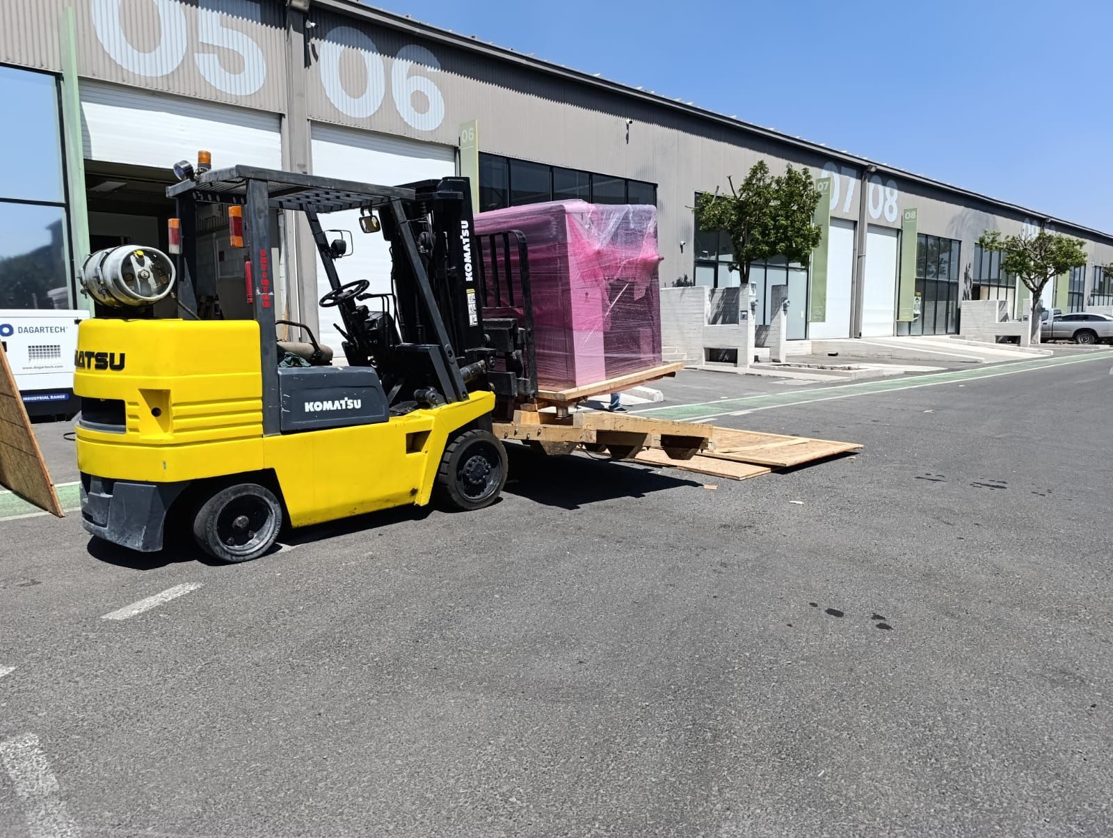 Una carretilla elevadora amarilla está cargando una caja rosa en un palé frente a un edificio.
