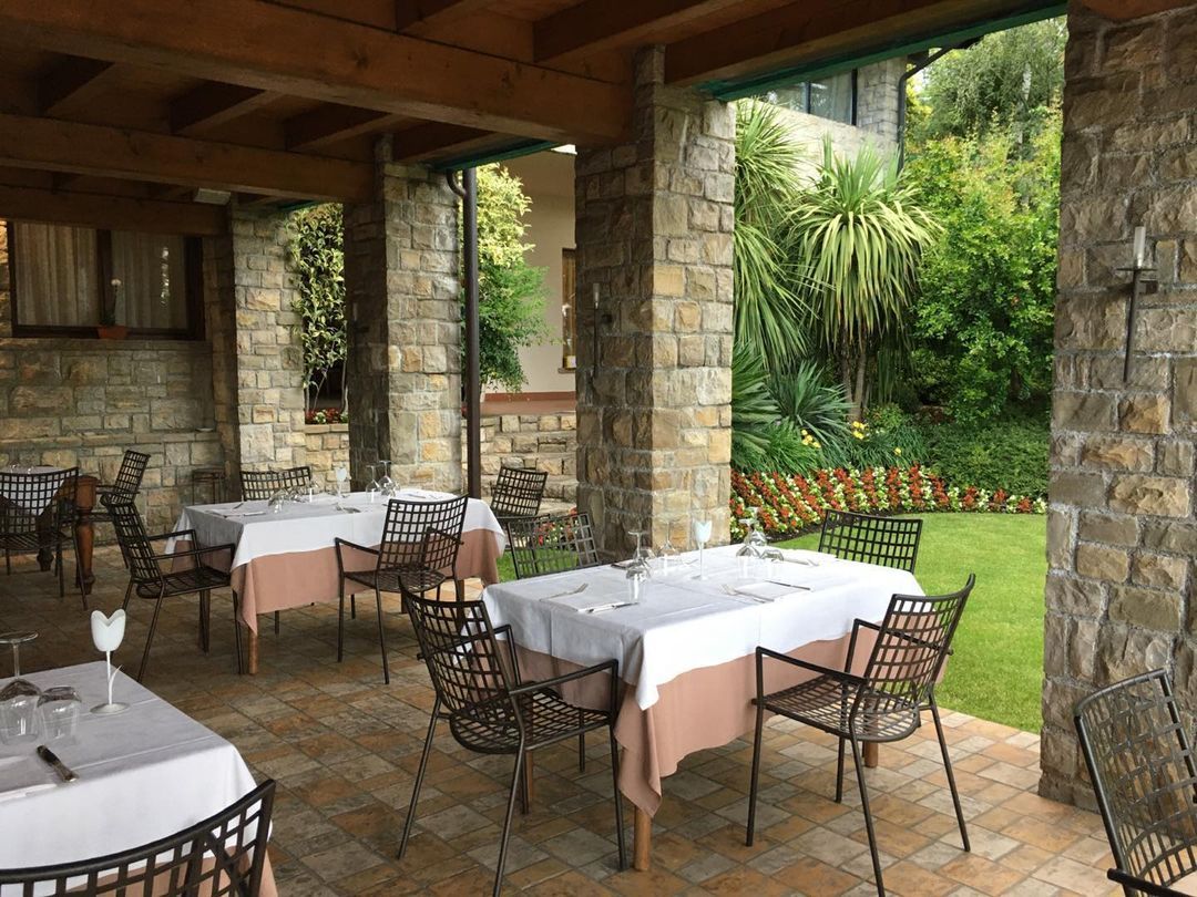 Zona pranzo all'aperto con colonne in pietra, tovaglie bianche e sedie apparecchiate per il pasto, con vista su un prato verde.