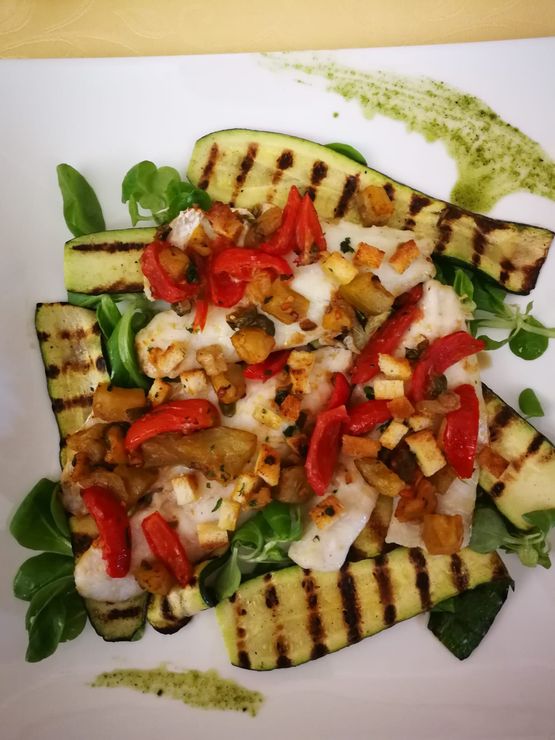Pesce alla griglia con zucchine, peperoni rossi, crostini e pesto su un piatto bianco.