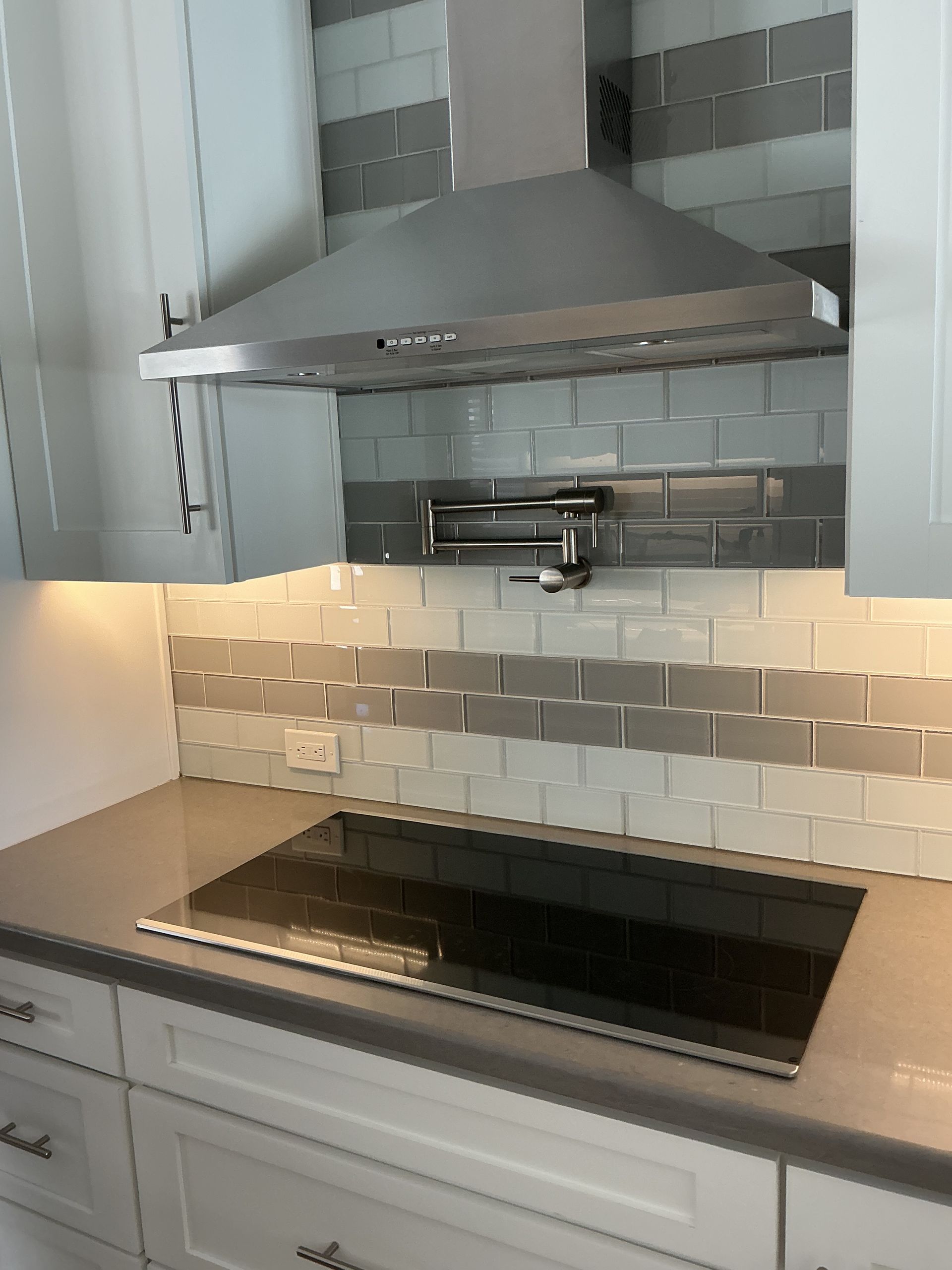 Modern kitchen with stainless steel range hood, cooktop, and gray and white tiled backsplash.