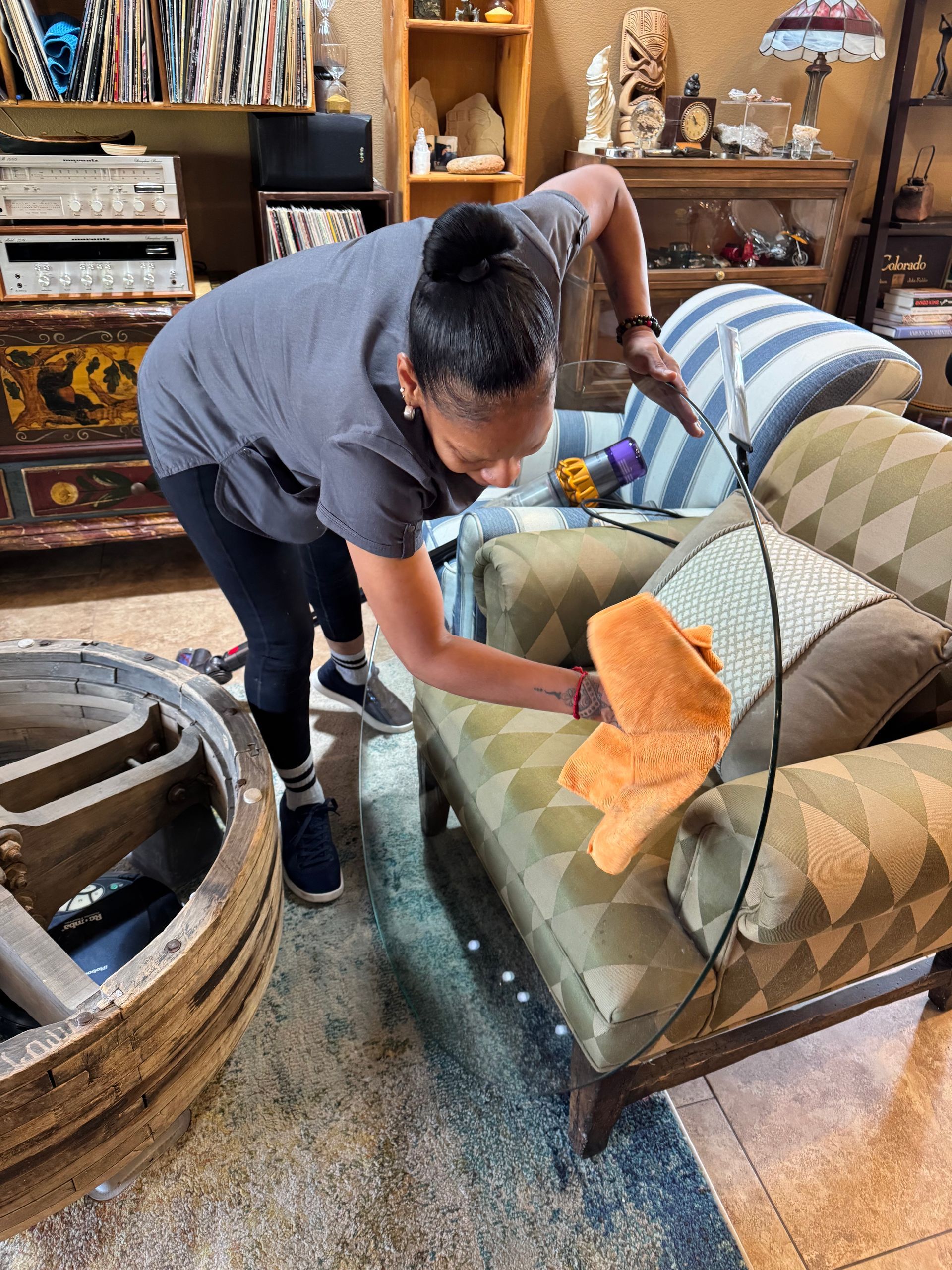 Woman dusting a patterned sofa in a shop filled with antique furniture and decor.