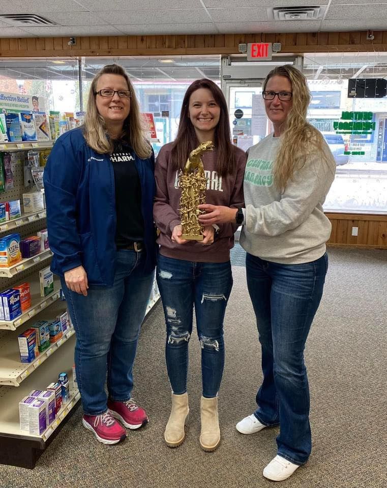 Three women are standing next to each other in a pharmacy holding a saxophone.