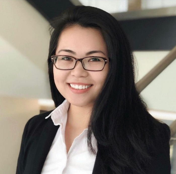 Woman with long black hair, glasses, smiling, wearing a blazer, in a modern building.