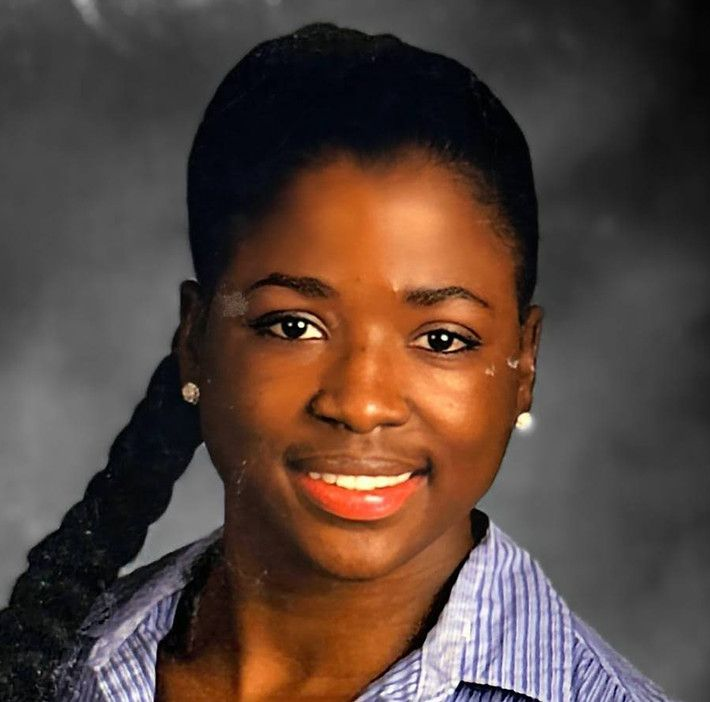 Person with dark skin and a braid smiles, wearing a striped blue shirt and pearl earrings.