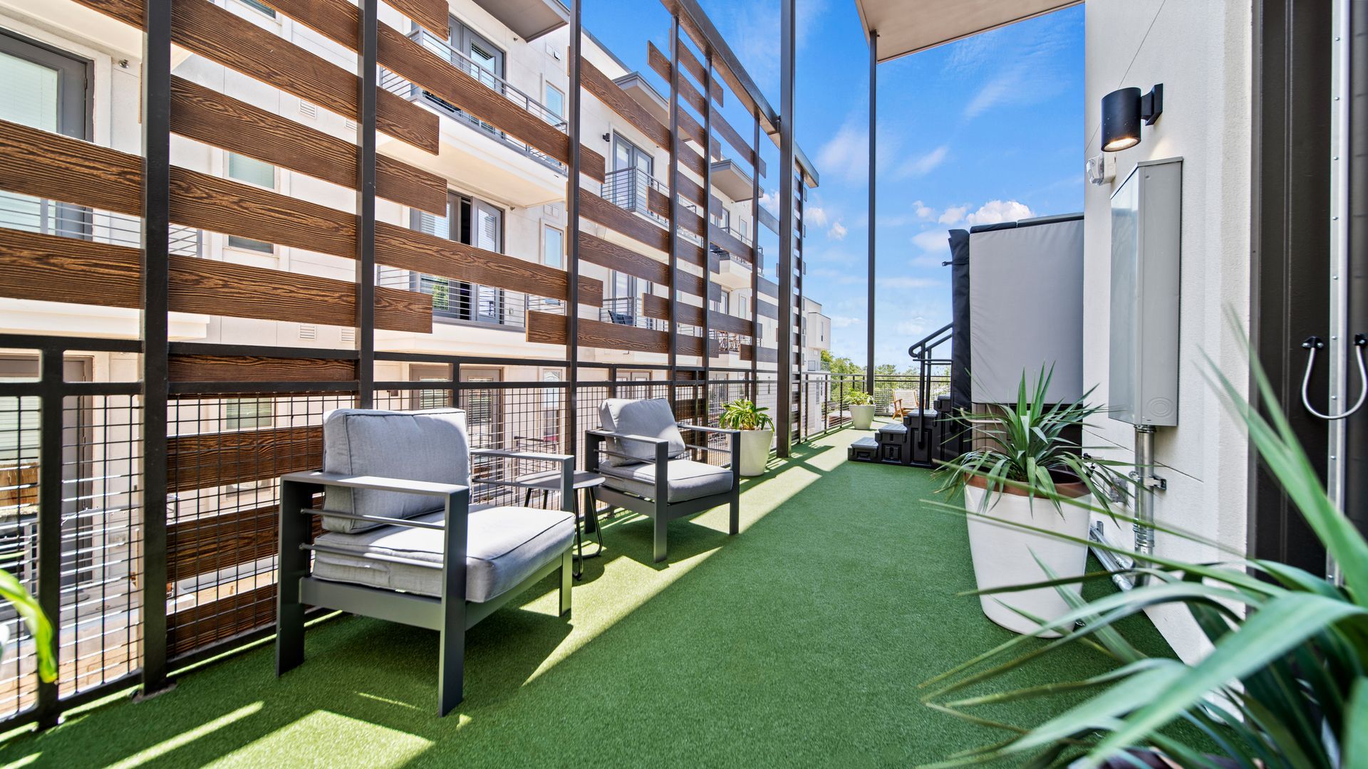 Apartment balcony with green turf, outdoor seating, and potted plants beside neighboring buildings
