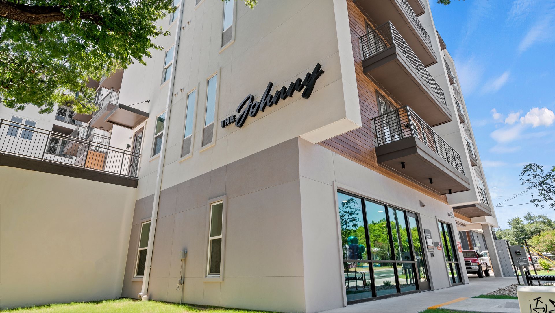 Modern white corner building with balconies, glass storefront windows, and a leafy tree overhead