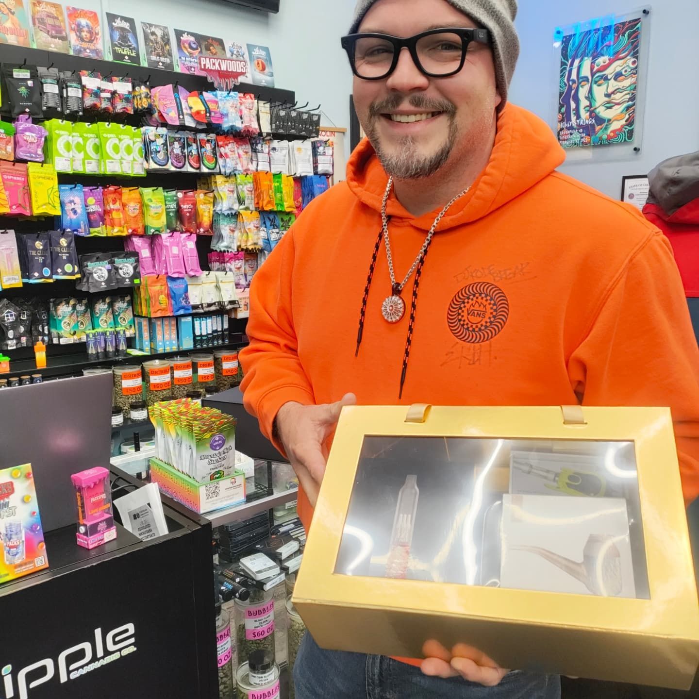Man in orange hoodie holding a golden box with a display case in a store. Shelves of products behind him.