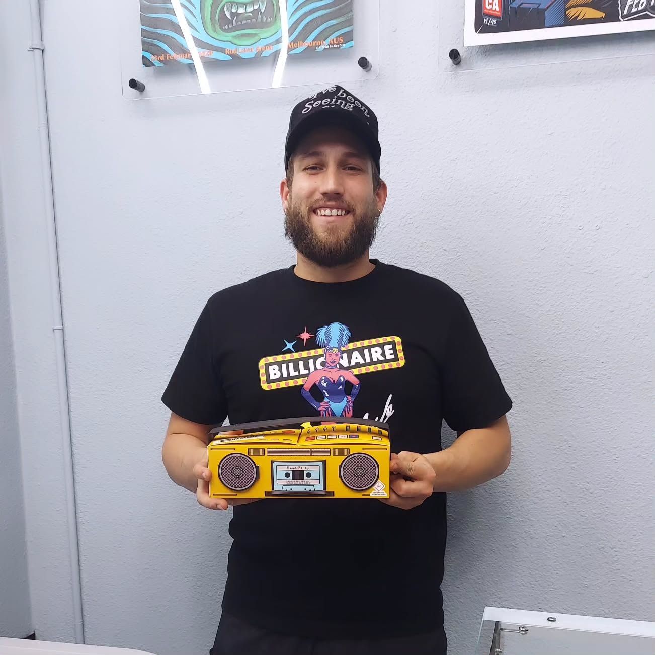Man holding yellow boombox-shaped object, smiling. Black t-shirt, cap, indoor setting with artwork.