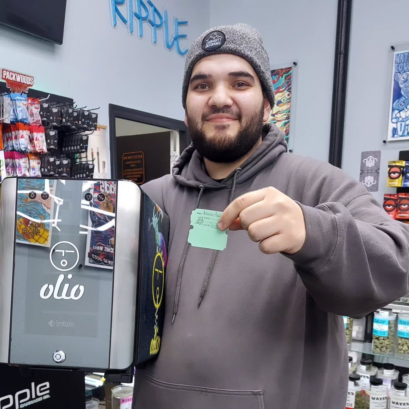 Man in gray hoodie and beanie holding a green tag near an oil machine in a store.