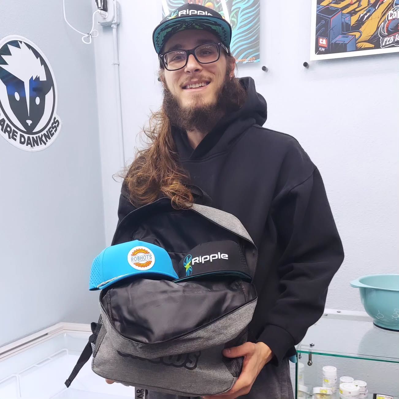 Man smiles, holding open a backpack with two caps inside; shop setting, logo on wall.