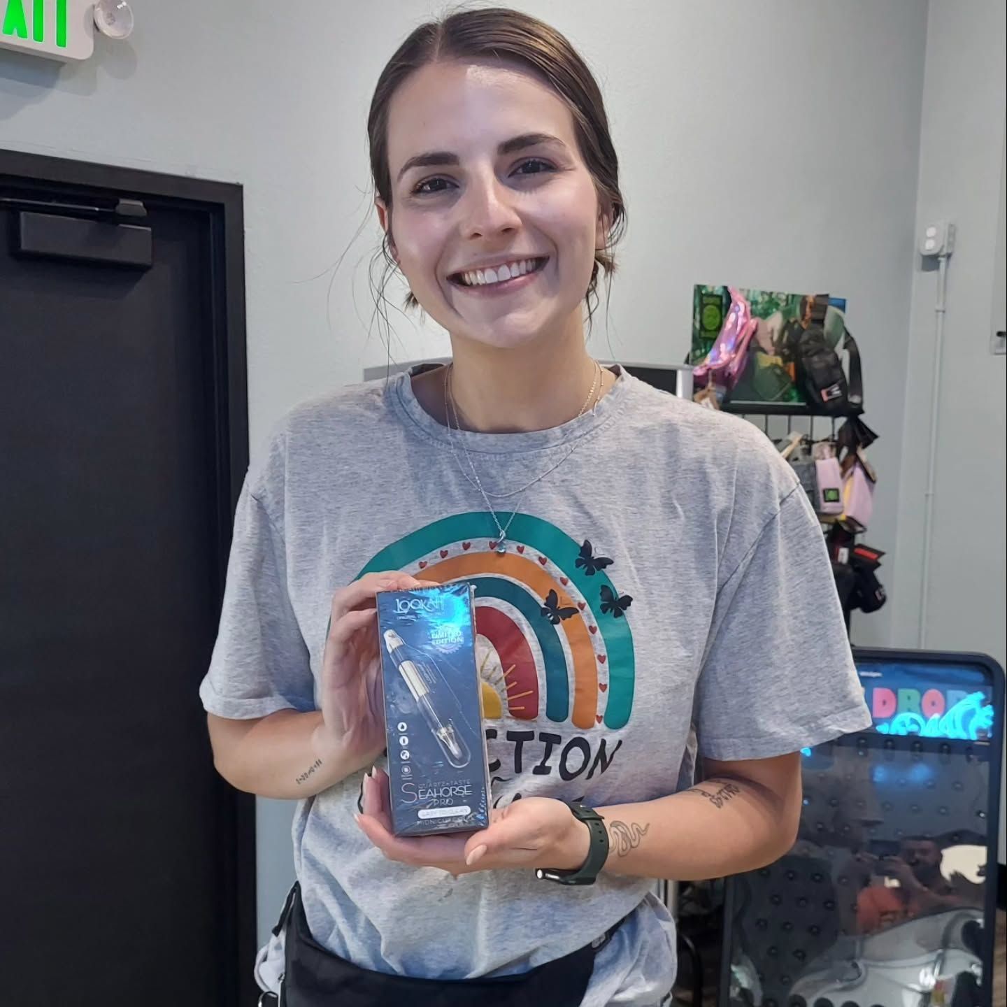 Woman holding a phone case, smiling. Gray t-shirt with a rainbow design, inside a shop.