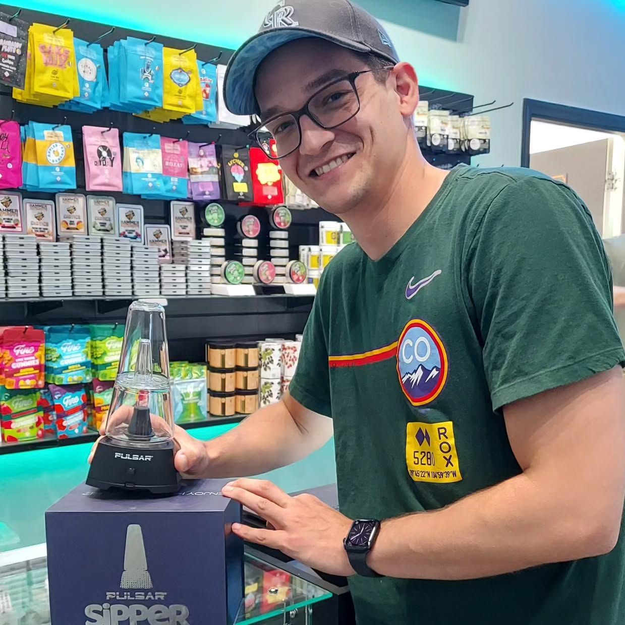 Man smiling, holding a black vaporizer, in a store with product displays.