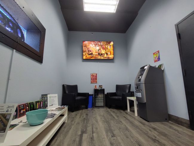 Interior of a waiting room with a TV, chairs, an ATM, and a low table with items on it.