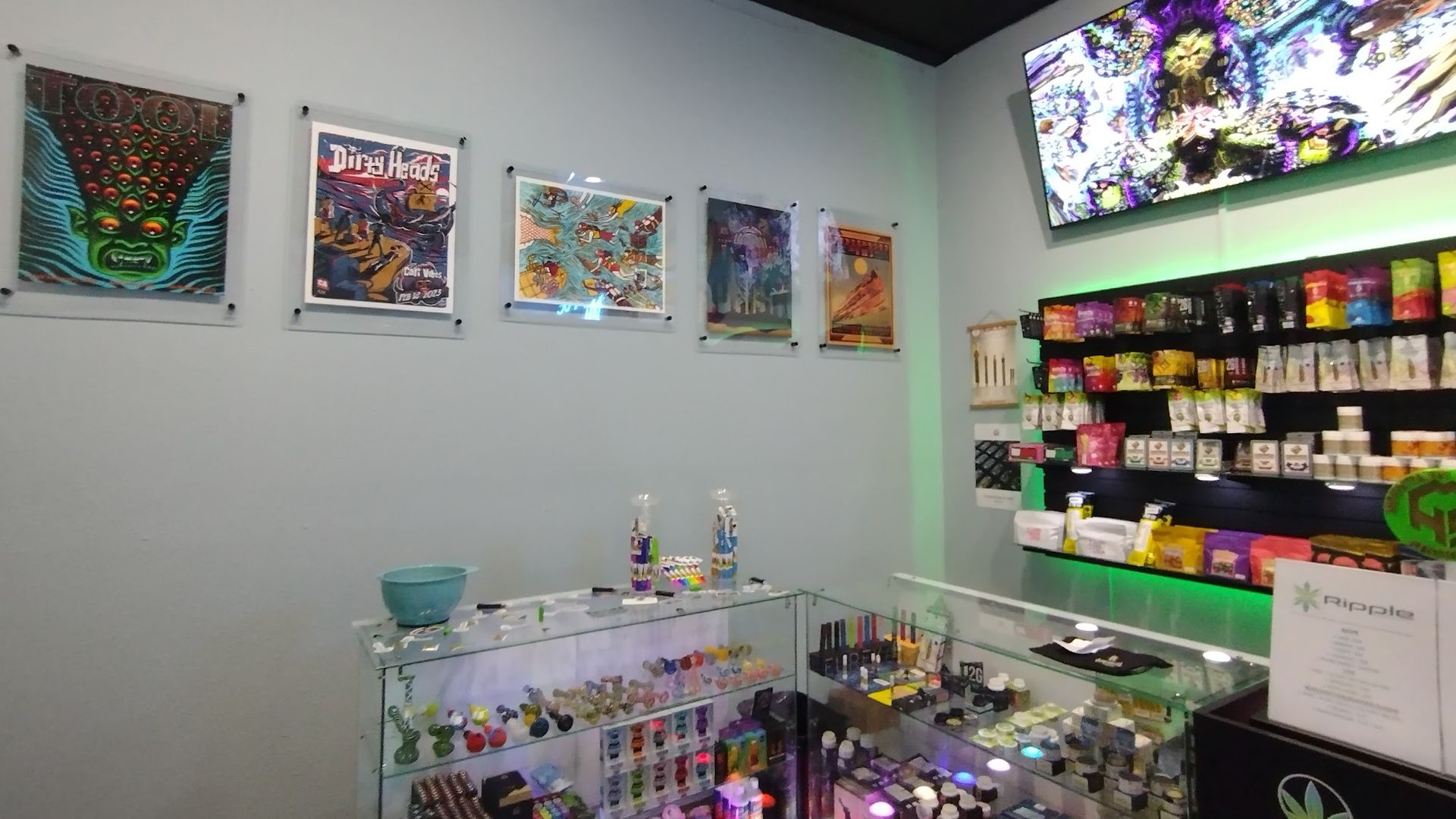 Inside a vape shop, art on the wall, lit display cases filled with products, a TV showing graphics.