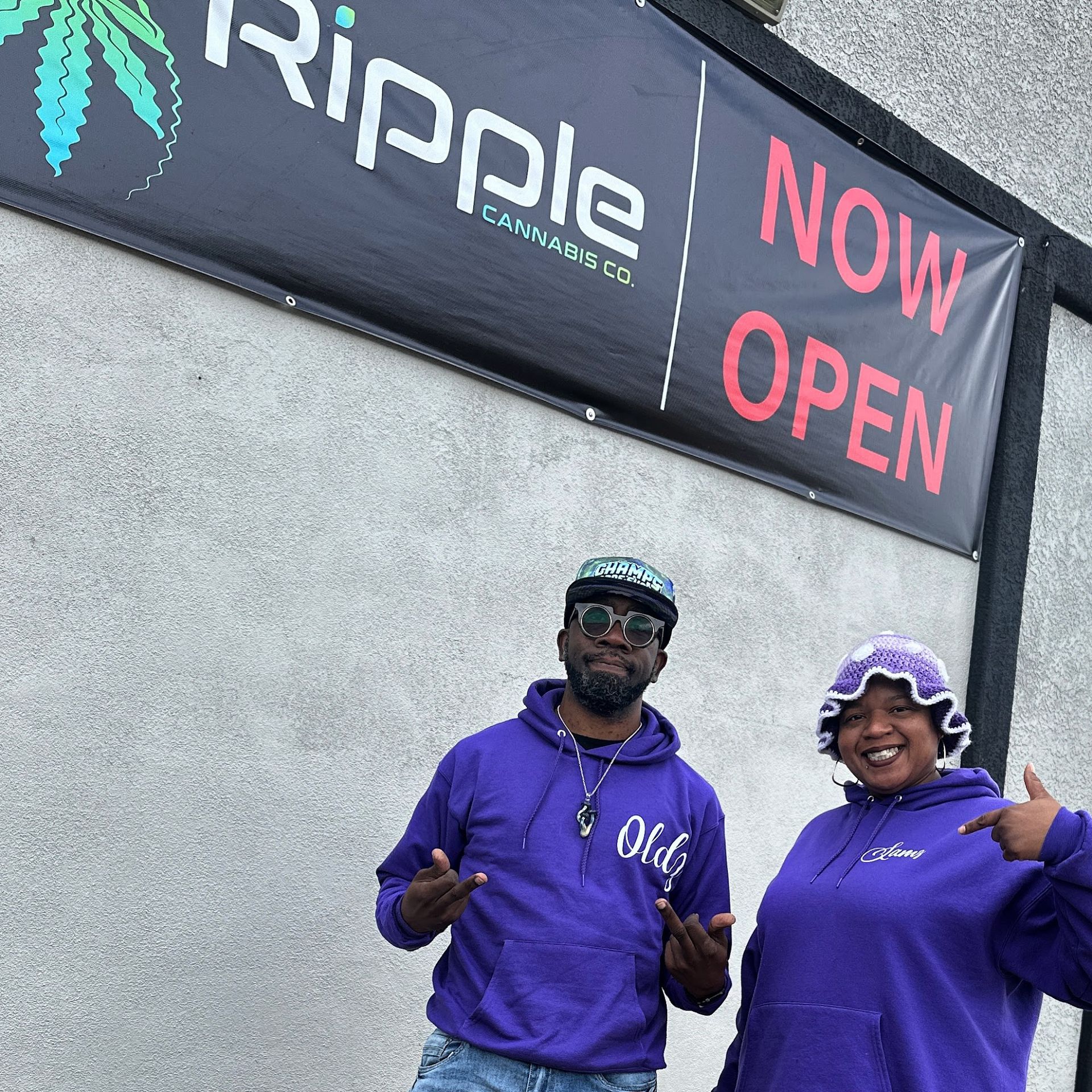 Two people in purple hoodies stand in front of a Ripple Cannabis Co. sign. 