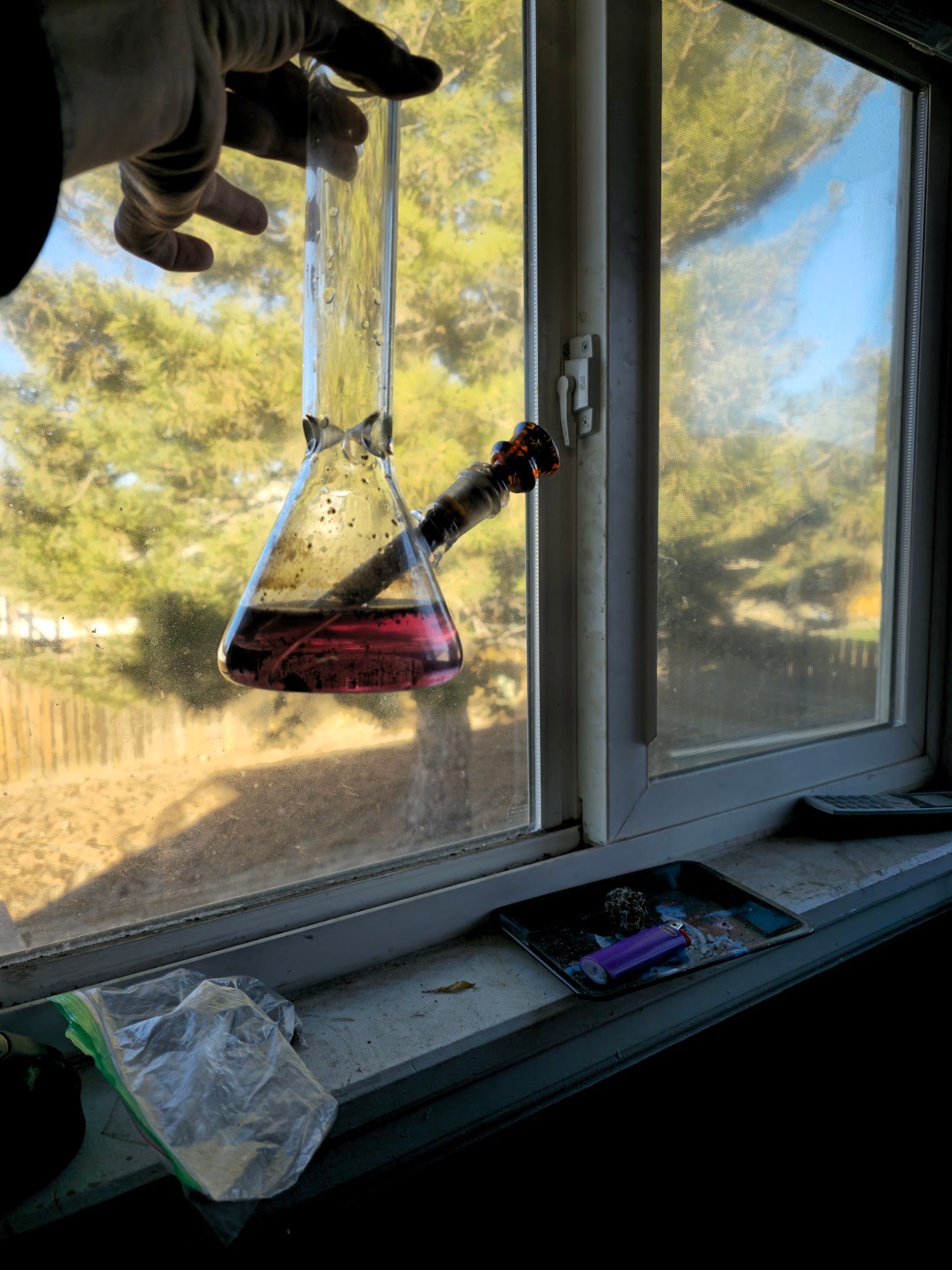 A bong held in front of a window, with pink liquid inside. A hand holds the glass.