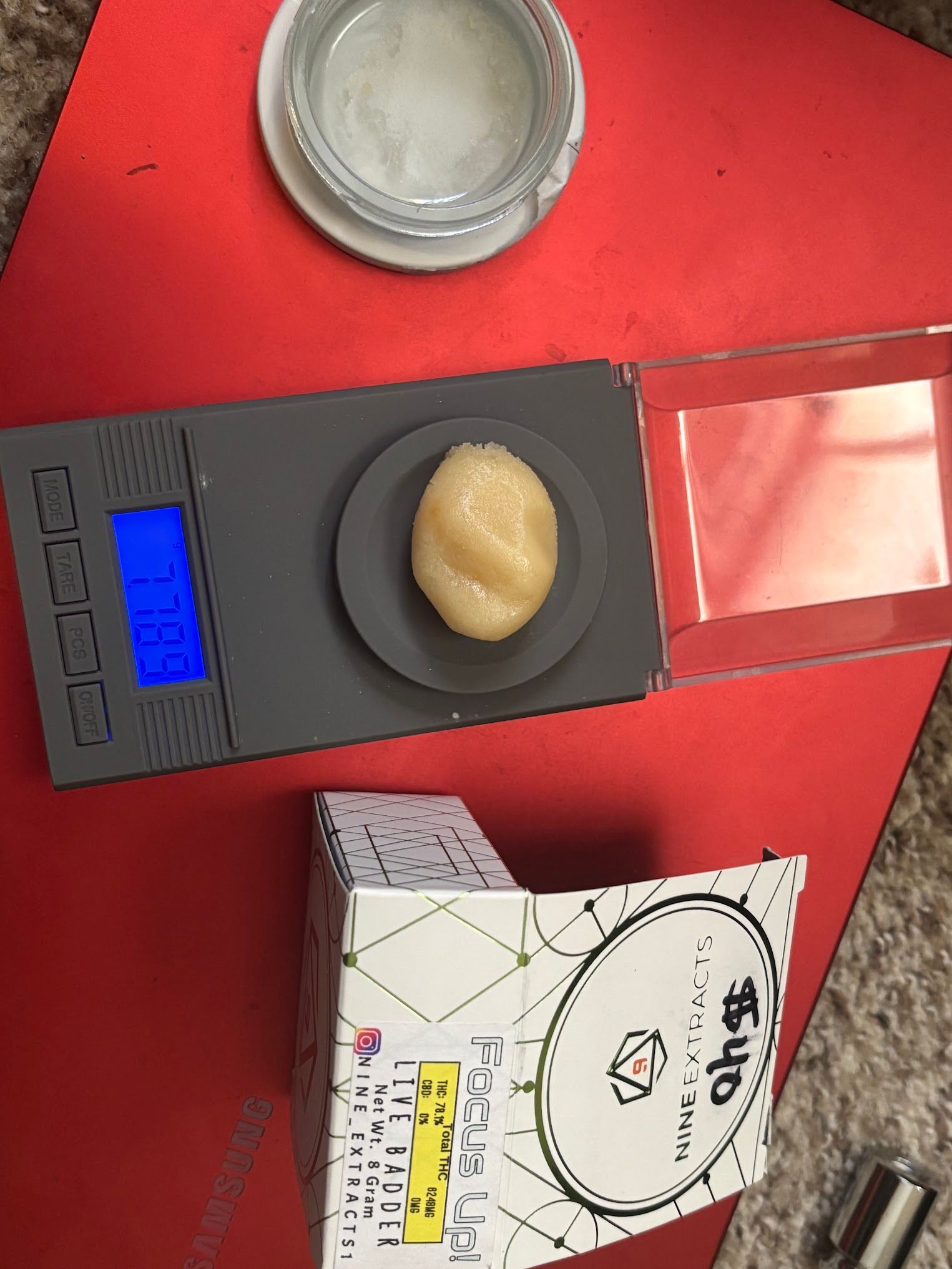 A scale with a small, round, light-colored substance on it. A container and a box are also present.