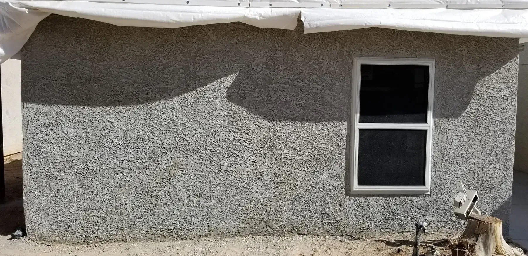 Exterior of a building with a textured gray stucco wall, a window, and a white covering over the roof.