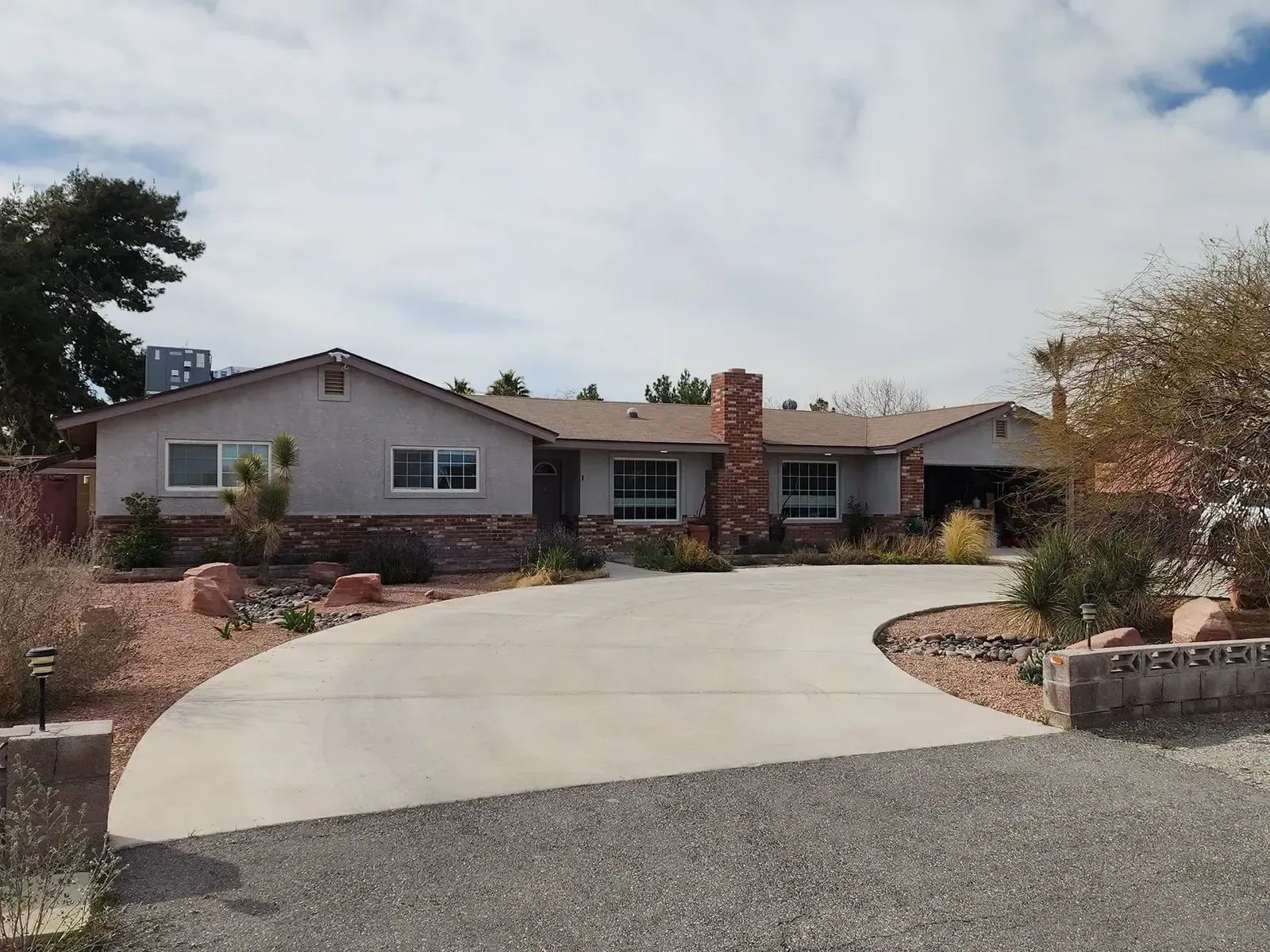 Ranch-style house with gray siding, brick accents, and a curved concrete driveway on a sunny day.