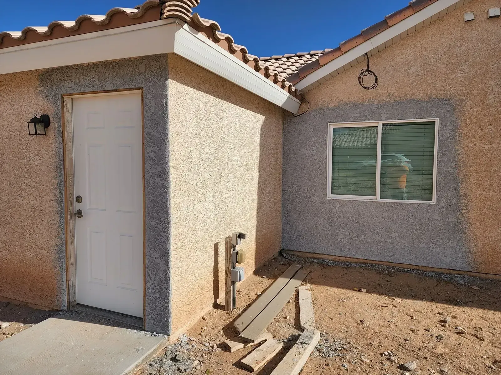 Exterior corner of a building, beige stucco with gray trim around a white door and window, construction debris nearby.