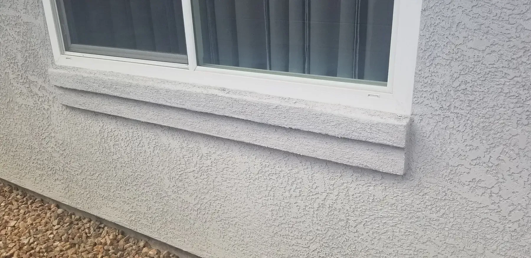 Exterior window with white trim, and a decorative ledge, set in a stucco wall.