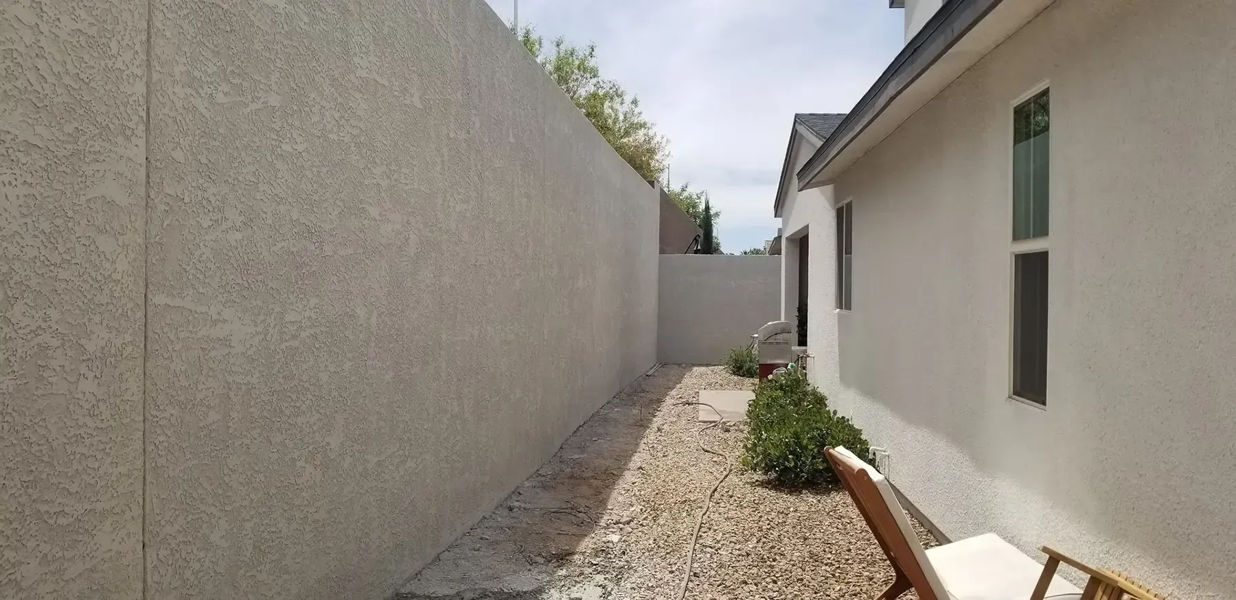 Narrow outdoor space with a gravel path, concrete walls, and a white house with windows.
