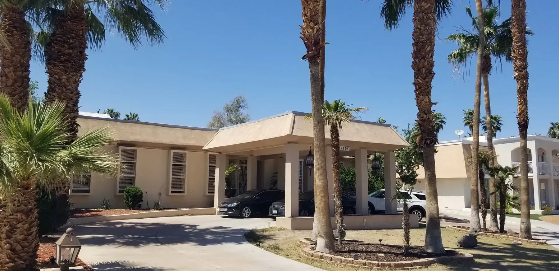 Tan building with covered entrance and palm trees under a blue sky.