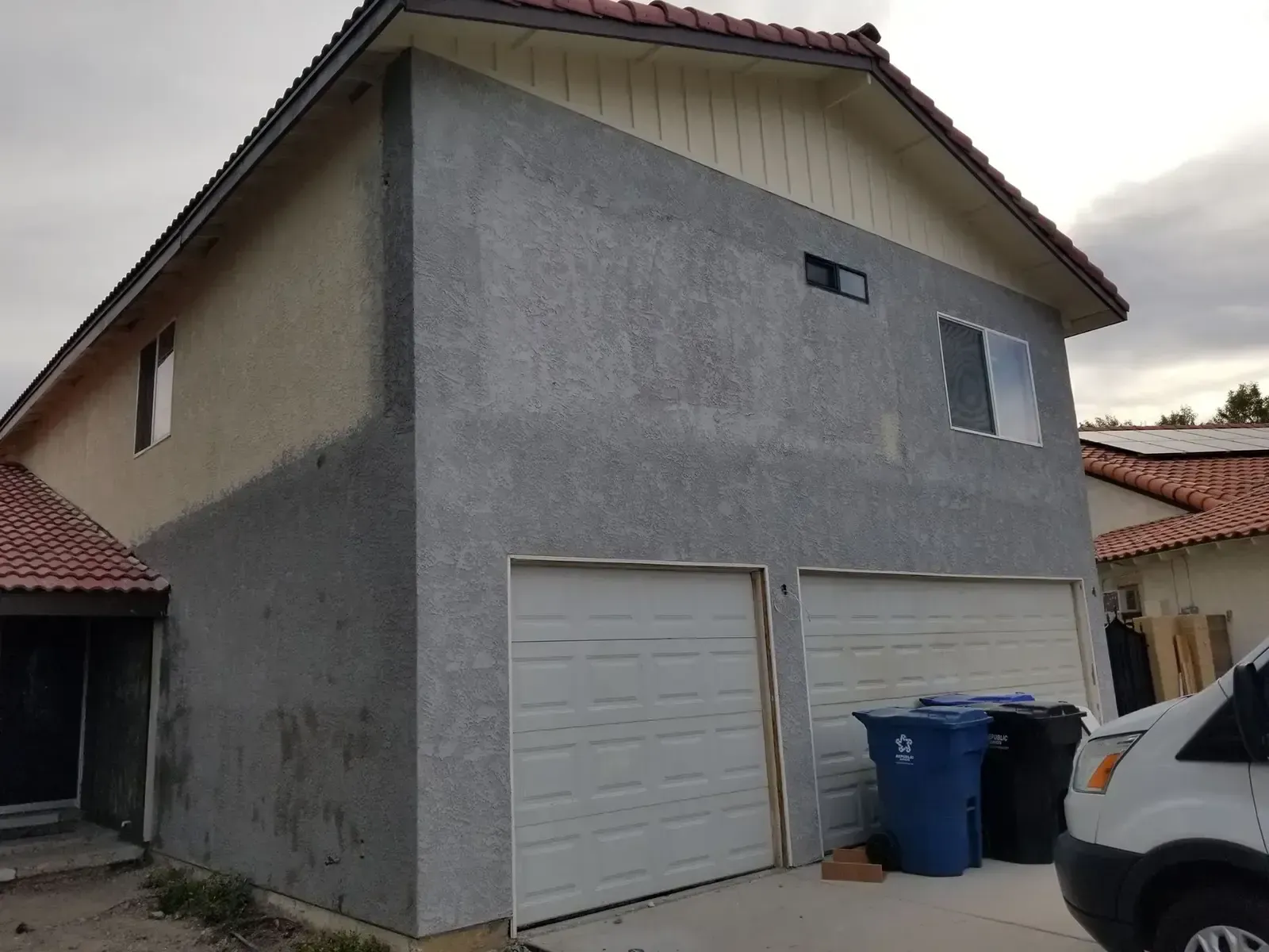Two-story house with tan and gray stucco, two garage doors, and a cloudy sky.