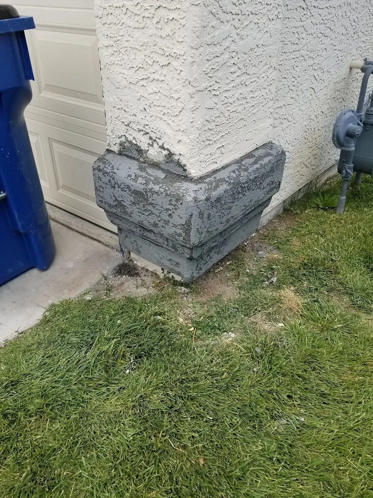 Corner of a stucco building with a gray, decorative base near grass and a blue trash can.