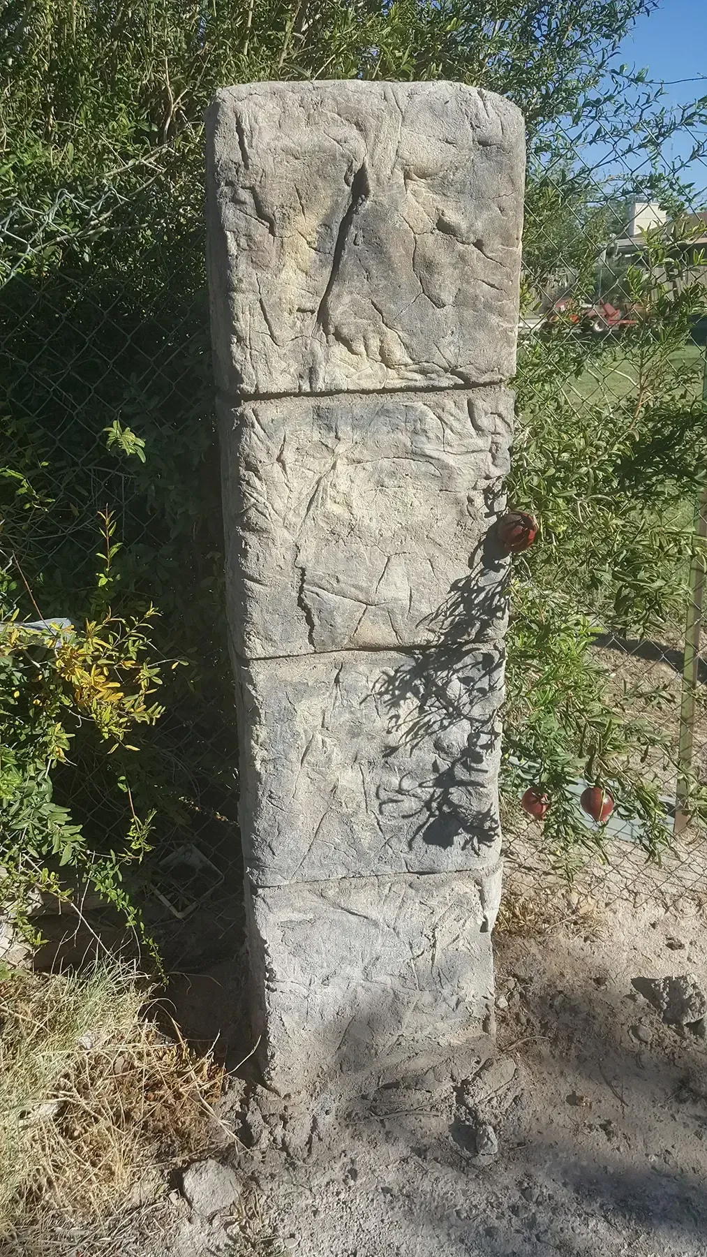 Stone pillar with three sections, beside a chain-link fence and foliage.