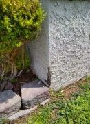 Damaged stucco wall next to a green bush, with cracked stone landscaping border.