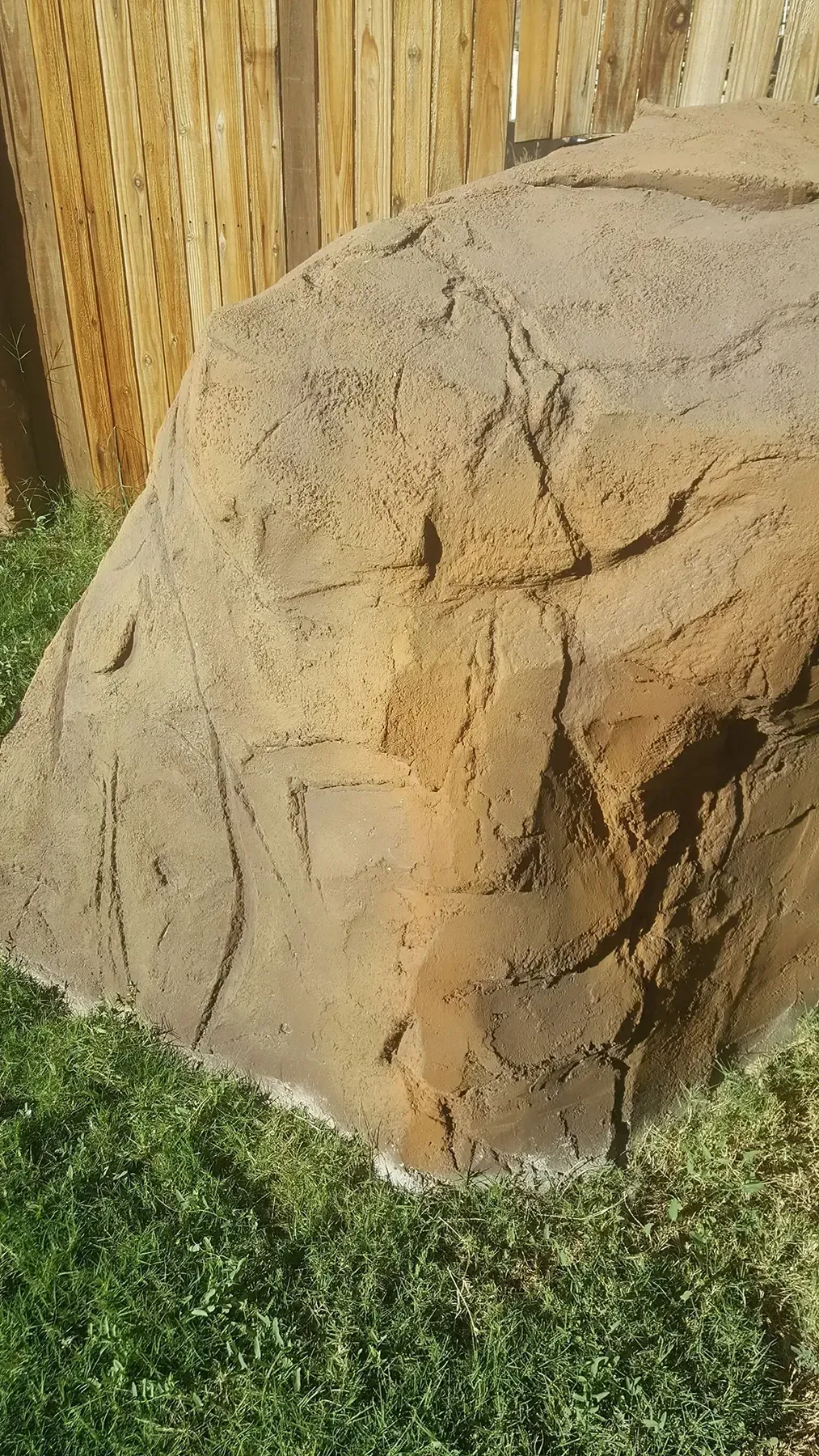 Large faux rock in a grassy yard, beside a wooden fence.