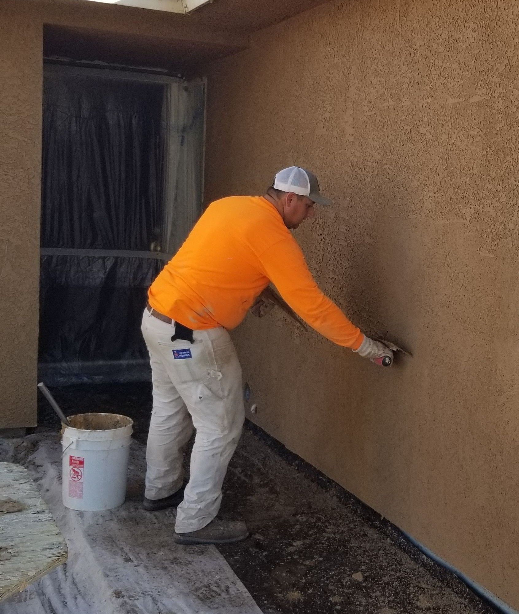 Person applying stucco to a wall outdoors, wearing orange shirt and white pants.