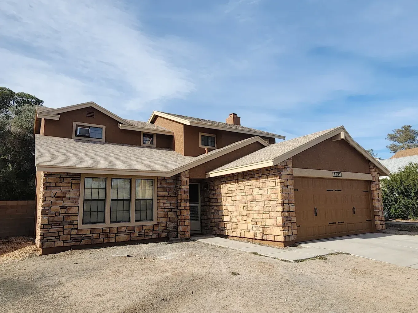 Two-story house with stone facade, tan stucco, and attached garage under a cloudy blue sky.