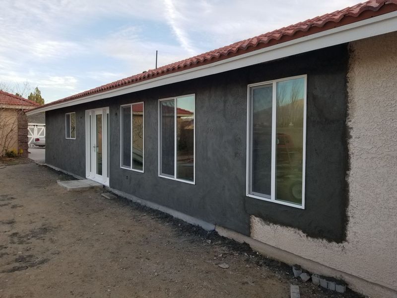 Exterior of a house with dark gray stucco siding, windows, and red tile roof, and partial unfinished beige stucco.