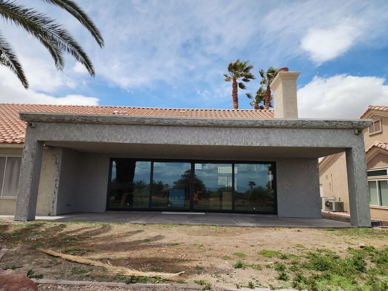 Rear view of a home's patio with large sliding glass doors, stucco walls, and a tiled roof, under a cloudy sky.