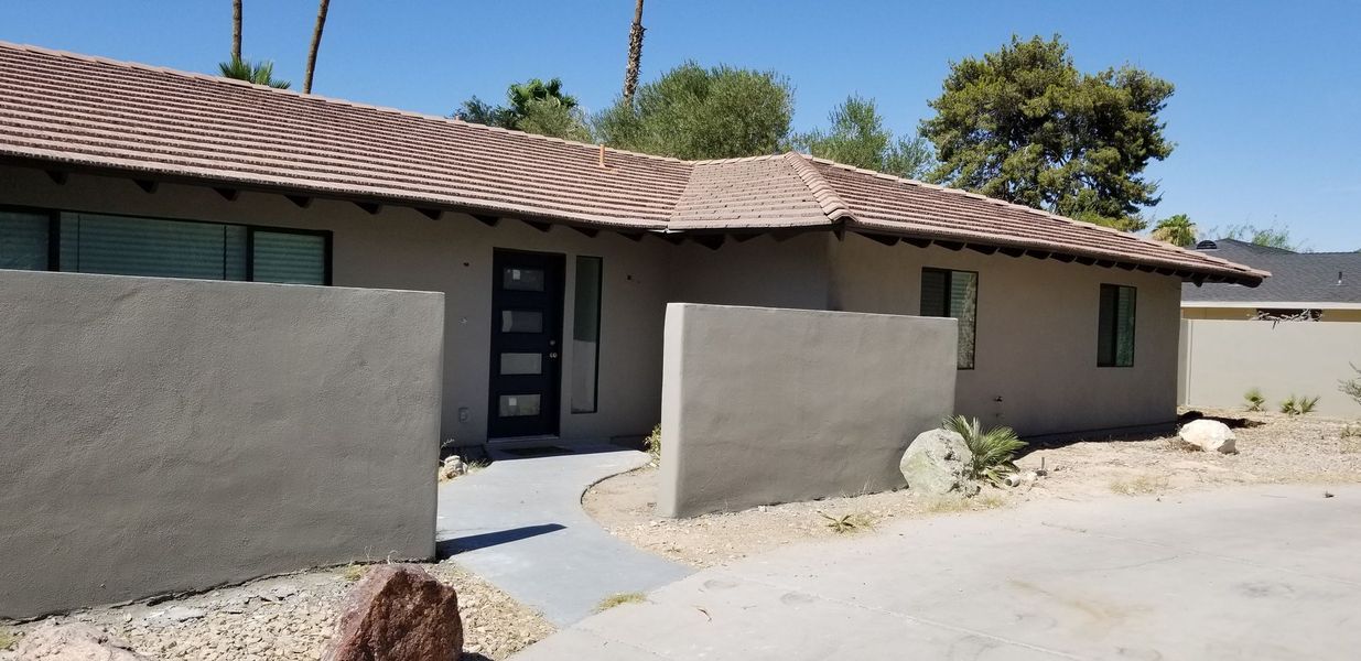 Exterior view of a single-story house with a brown tile roof and gray walls, a walkway, and desert landscaping.
