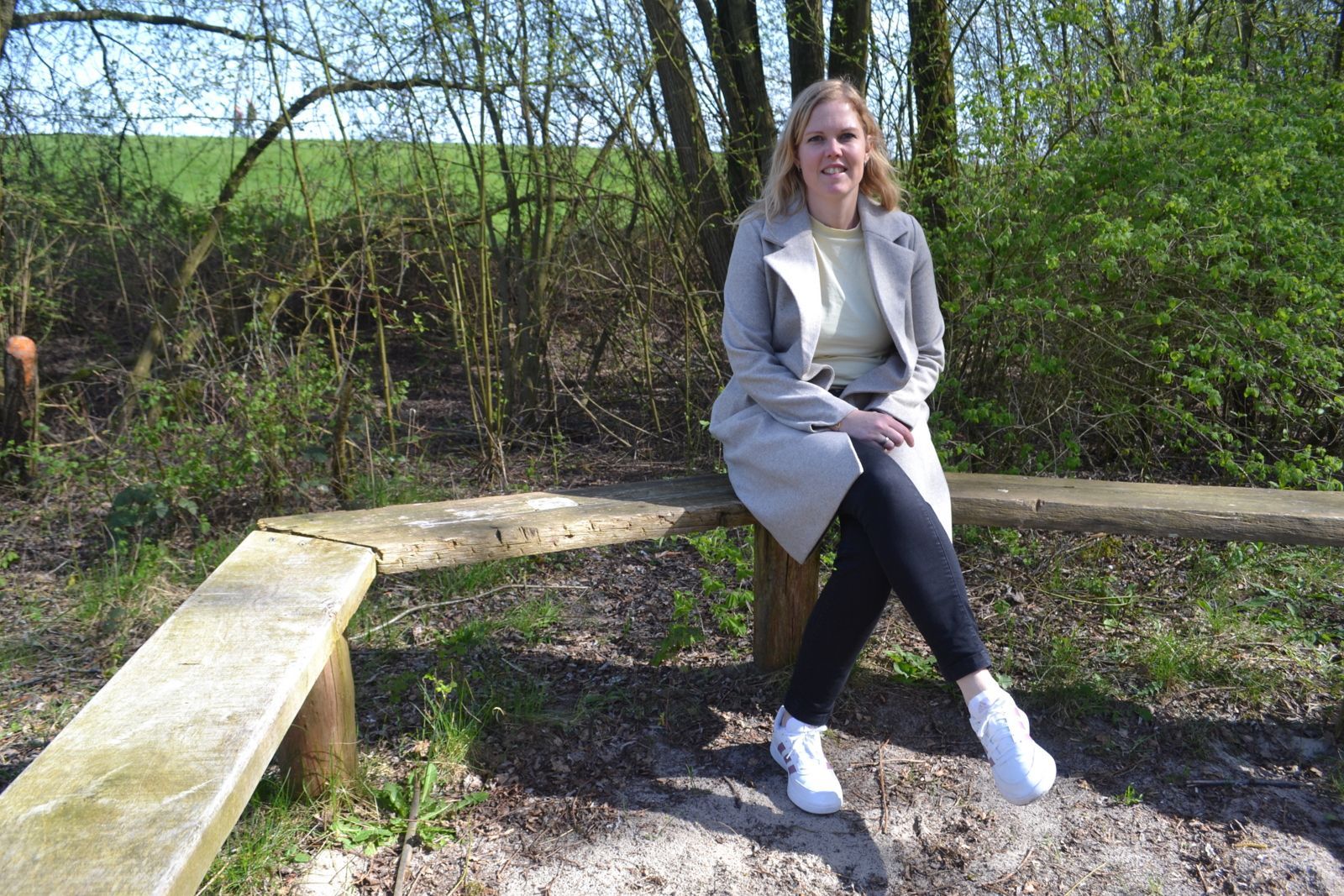 Een glimlachend persoon in een lichte jas en donkere legging zit op een L-vormige houten bank in een bosrijk, grasrijk buitengebied.