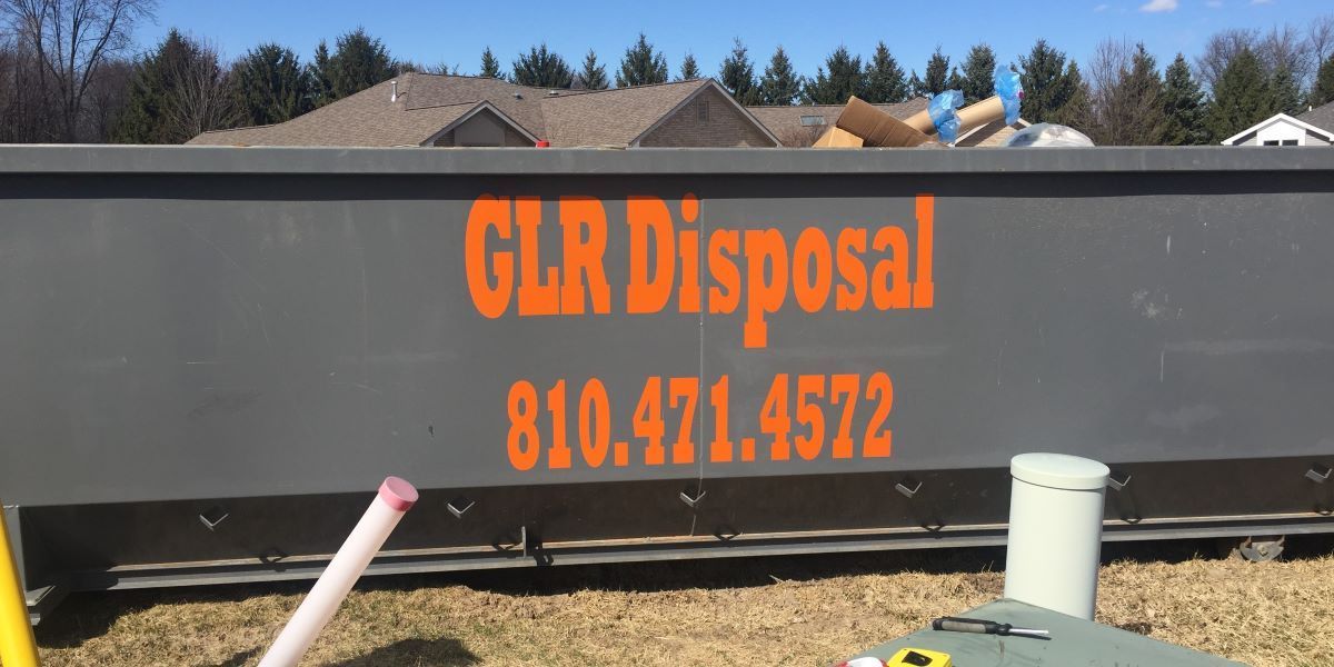 A gray dumpster from Great Lakes Region Disposal, LLC, with phone number 810.471.4572, sits on grass before a rustic barn.
