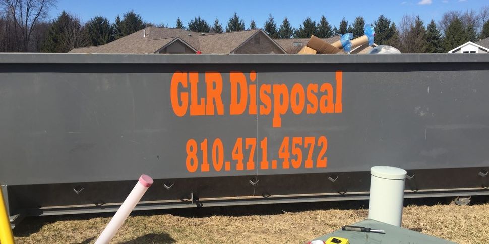A gray dumpster from Great Lakes Region Disposal, LLC, with phone number 810.471.4572, sits on grass before a rustic barn.