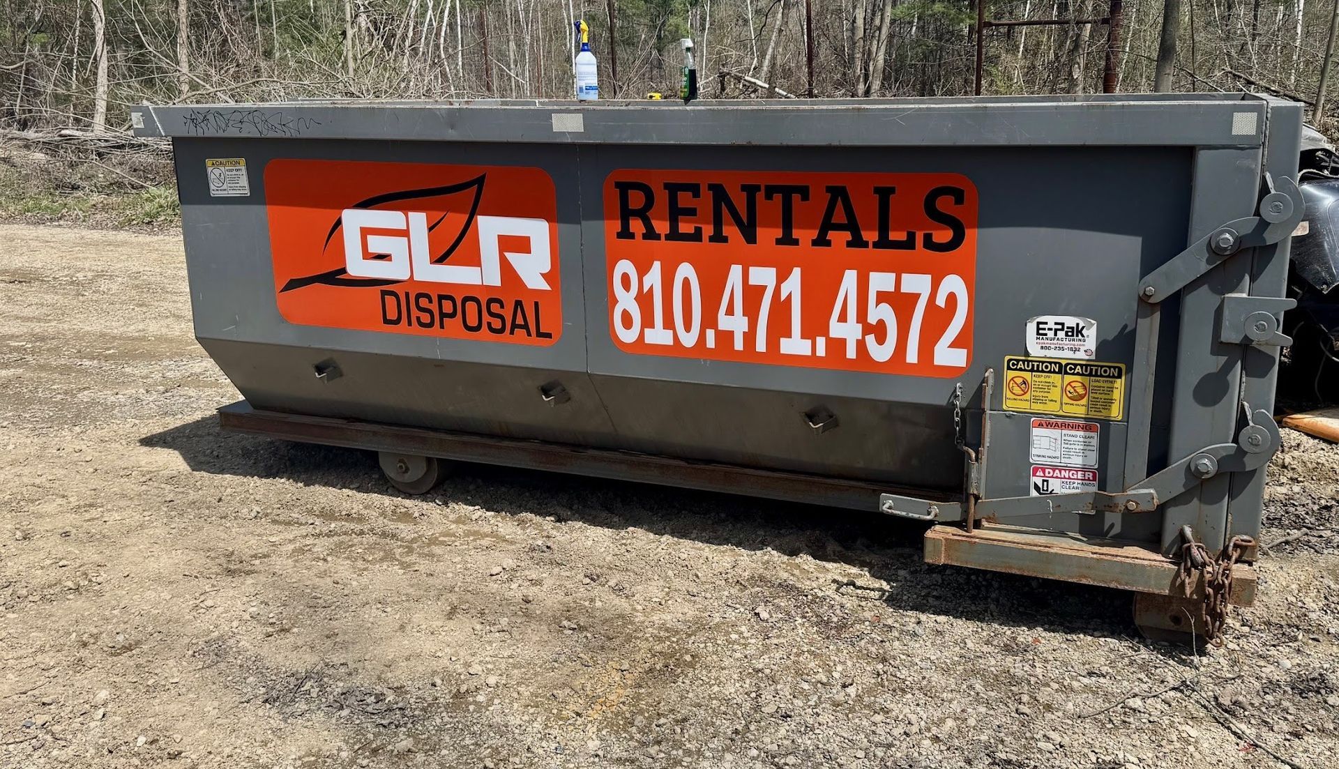 A grey roll-off dumpster with orange branding for GLR Disposal and a rental phone number on the side, sitting on dirt.